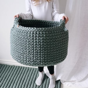 May include: A large, round, sage green storage basket crafted from thick, chunky knit cord. The basket features two integrated handles on the sides and a small, light brown leather-like tag attached to the upper rim of the textured, woven structure.