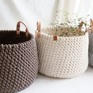 Könnte beinhalten: Drei handgefertigte, runde Aufbewahrungskörbe in Braun-, Creme- und Grautönen. Jeder Korb hat ein dickes, strukturiertes Strickdesign und Ledergriffe, die mit Metallnieten befestigt sind. Ein Korb enthält Grünzeug.