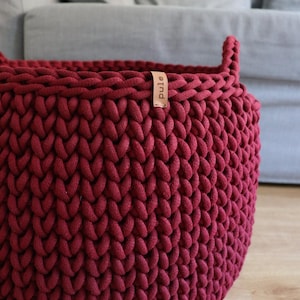 May include: A burgundy, hand-knitted basket with a sturdy, rounded shape. The basket is made of thick, textured yarn, with a small leather tag that reads "pule". The basket has two handles and is sitting on a wooden floor.