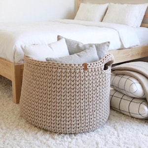 Op de afbeelding: Een grote, ronde opbergmand van beige, grof gebreid garen. De mand is voorzien van geïntegreerde handgrepen aan de zijkanten en een klein, rechthoekig bruin label in lederlook aan de bovenrand. De mand is gevuld met verschillende zachte kussens.