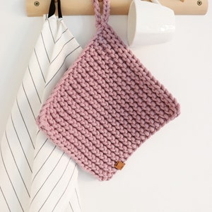 Op de afbeelding: Een vierkante, gehaakte pannenlap in stoffig roze, met een ophanglus. Hij hangt aan een houten rek met een gestreepte theedoek en een witte mok. Afmetingen ongeveer 20 cm.