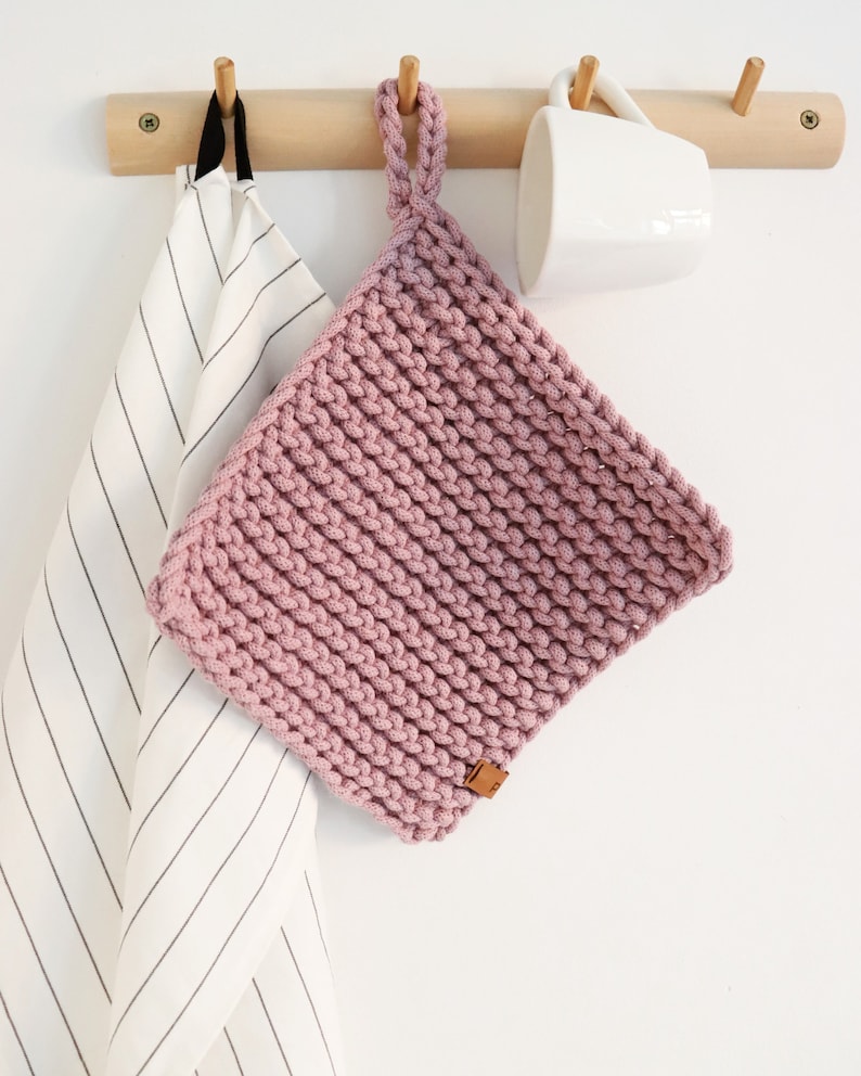 Op de afbeelding: Een vierkante, gehaakte pannenlap in stoffig roze, met een ophanglus. Hij hangt aan een houten rek met een gestreepte theedoek en een witte mok. Afmetingen ongeveer 20 cm.