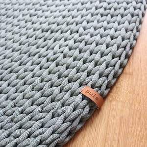 Könnte beinhalten: Ein runder, grauer, gestrickter Teppich mit einer dicken, geflochtenen Textur. Ein kleines, braunes Lederetikett mit dem Wort "pule" ist am Rand befestigt. Der Teppich besteht aus einem weichen, strapazierfähigen Material.
