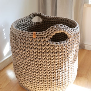 May include: A large, round, taupe-colored crochet basket with two integrated handles. The basket has a thick, textured appearance and a small leather tag with the word "pule" on it. The basket is sitting on a wooden floor.