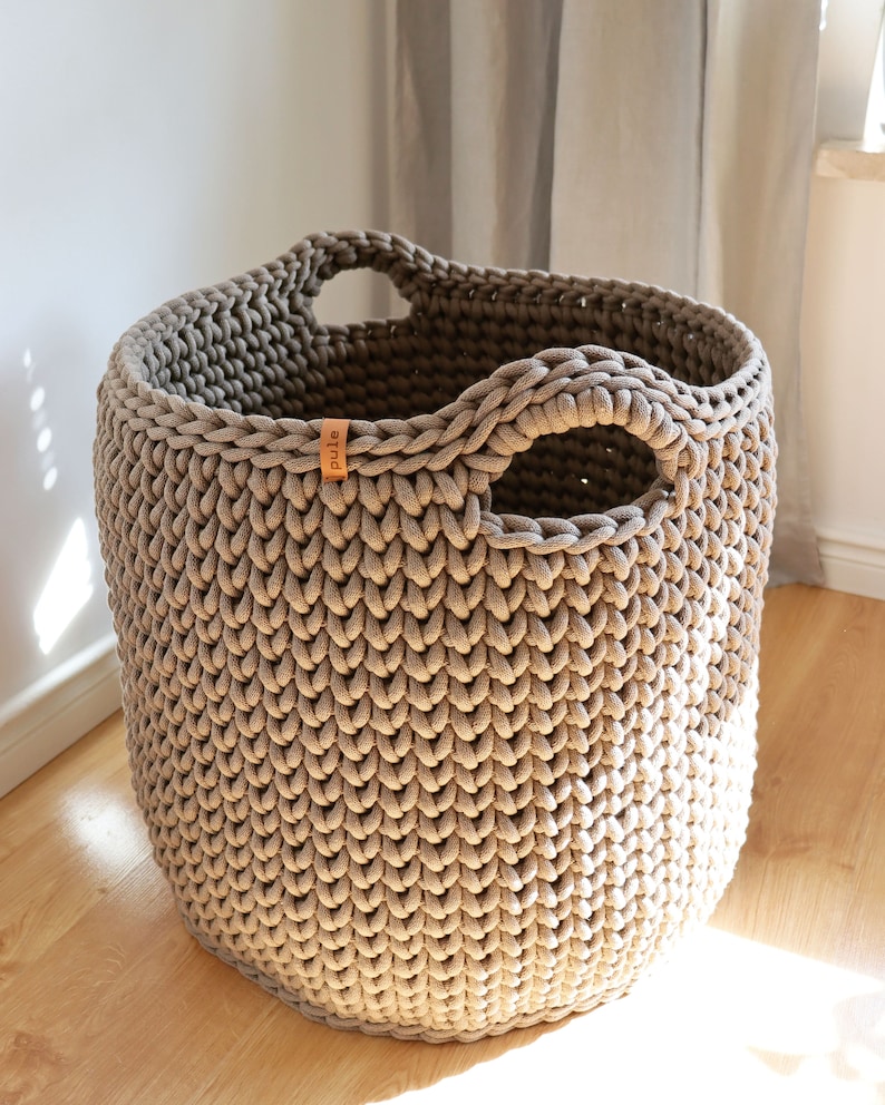 May include: A large, round, taupe-colored crochet basket with two integrated handles. The basket has a thick, textured appearance and a small leather tag with the word "pule" on it. The basket is sitting on a wooden floor.