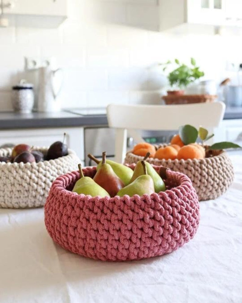 May include: Three handmade crochet baskets filled with fruit. The front basket is a dusty rose color and holds green pears. The other two baskets are beige and hold plums and oranges. The baskets are on a white tablecloth.