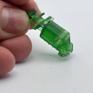 以下が含まれることがあります： 幾何学的なデザインの半透明の緑色のプラスチック製の物体。白い背景に対して、人の指で物体が保持されています。長方形の本体には一連の突起があり、小さな濃い緑色のキャップが付いています。