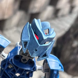 May include: A blue plastic action figure head featuring a textured, angular mask with sharp geometric lines and prominent eye slits. Bright red eyes are visible through the openings of the mask, which is attached to a blue and gray articulated torso.