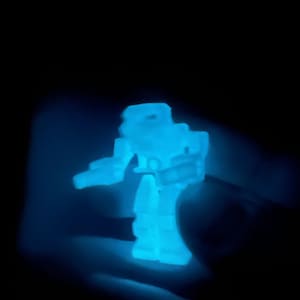 May include: A blue glow-in-the-dark plastic toy robot. The robot is standing on a hand.