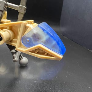 以下が含まれることがあります： ベージュとブルーの玩具ロボット。透明なブルーの頭と回転する腕が2本あります。ロボットは、歯車や関節が見える詳細なデザインです。