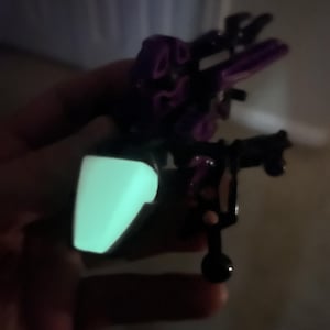 以下が含まれることがあります： 胸に緑色の光が輝く黒と紫のロボットのおもちゃ。ロボットは、複数の関節と腕の先端に小さなボールが付いた、詳細なデザインです。