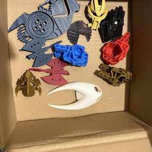 May include: A collection of 3D printed plastic toys in a cardboard box. The toys are in various colors, including gold, red, blue, white, and black. Some of the toys are shaped like robots, while others are more abstract.