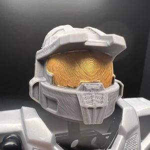 May include: A grey plastic 3D printed helmet with a gold visor. The helmet has a textured surface and is designed to look like a futuristic soldier's helmet.