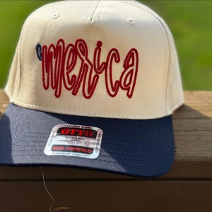 Könnte beinhalten: Cremefarbene Baseballkappe mit marineblauem Schirm. Das Wort "America" ist in roter Schrift auf die Vorderseite gestickt. Die Kappe hat eine klassische Passform und einen gebogenen Schirm.