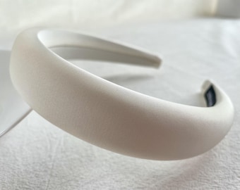 Könnte beinhalten: Ein weißes Satin-Haarband mit einem glatten, geschwungenen Design. Das Haarband ist dick und breit und perfekt, um jedem Outfit einen Hauch von Eleganz zu verleihen.