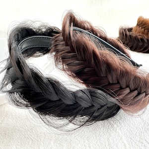 May include: Braided hair headbands in black and brown. The headbands feature a thick, woven braid design attached to a black headband base. A third headband in a lighter brown shade is visible in the background.