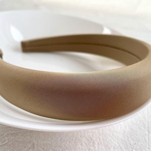 K&ouml;nnte beinhalten: Ein breites, beiges Stirnband aus weichem Stoff. Das Stirnband ist glatt und hat einen dezenten Glanz.