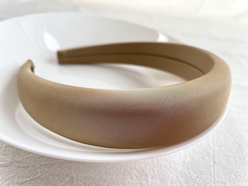 K&ouml;nnte beinhalten: Ein breites, beiges Stirnband aus weichem Stoff. Das Stirnband ist glatt und hat einen dezenten Glanz.