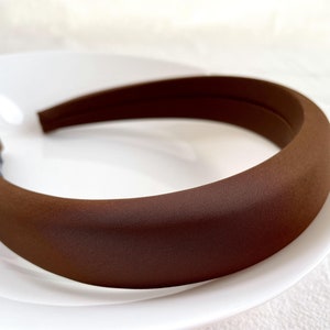 K&ouml;nnte beinhalten: Ein braunes gepolstertes Stirnband f&uuml;r Haare. Das Stirnband ist aus einem weichen, glatten Stoff.