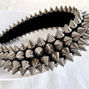 May include: A silver headband with a row of sharp, pointed spikes. The headband is made of a black fabric base and is covered in silver spikes.