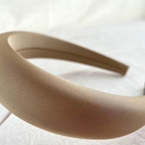 K&ouml;nnte beinhalten: Ein breites, beiges Stirnband aus weichem, glattem Stoff. Das Stirnband ist gebogen und hat einen leichten Glanz.