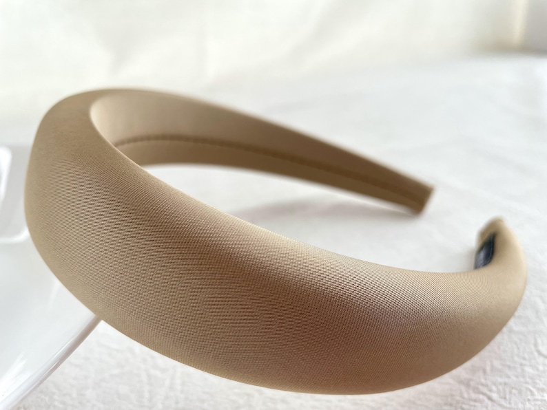K&ouml;nnte beinhalten: Ein breites, beiges Stirnband aus weichem, glattem Stoff. Das Stirnband ist gebogen und hat einen leichten Glanz.
