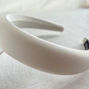 K&ouml;nnte beinhalten: Ein wei&szlig;es Satin-Haarband mit einem glatten, geschwungenen Design. Das Haarband ist dick und breit und perfekt, um jedem Outfit einen Hauch von Eleganz zu verleihen.