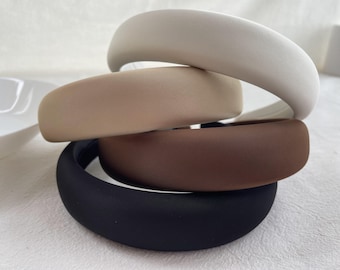 May include: Four fabric headbands stacked on top of each other. The top headband is white, the second is beige, the third is brown, and the bottom headband is black.