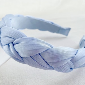 May include: A light blue braided headband made of soft fabric. The headband is a simple and elegant accessory for any occasion.