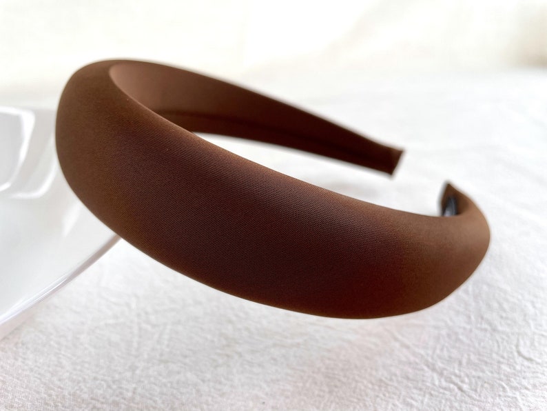 K&ouml;nnte beinhalten: Ein braunes gepolstertes Stirnband f&uuml;r Haare. Das Stirnband ist aus einem weichen, glatten Stoff.