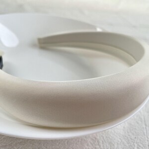 K&ouml;nnte beinhalten: Ein wei&szlig;es gepolstertes Stirnband f&uuml;r Haare. Das Stirnband ist aus einem weichen, glatten Material gefertigt und hat ein schlichtes, elegantes Design.