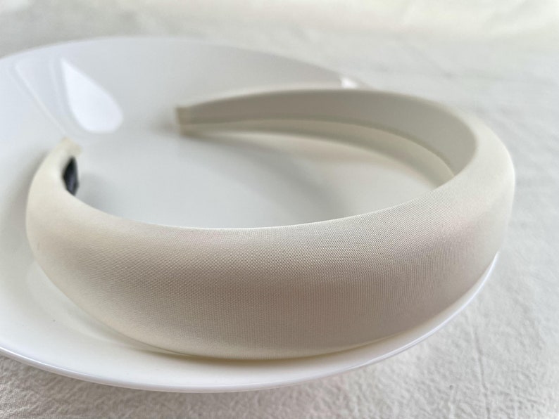 K&ouml;nnte beinhalten: Ein wei&szlig;es gepolstertes Stirnband f&uuml;r Haare. Das Stirnband ist aus einem weichen, glatten Material gefertigt und hat ein schlichtes, elegantes Design.
