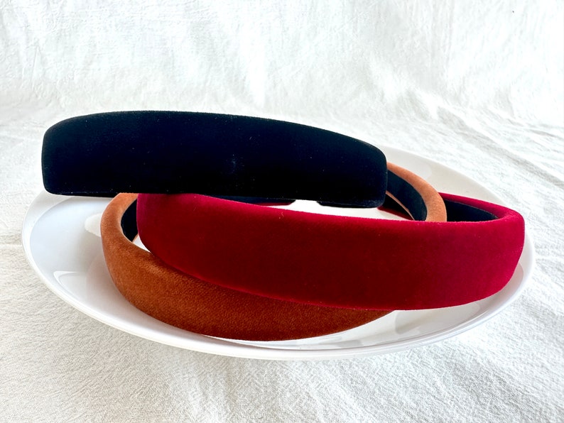 May include: Three velvet headbands in black, burgundy, and brown. The headbands are stacked on top of each other on a white plate.