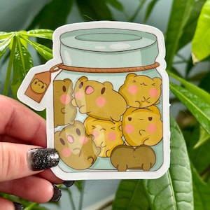 Pode incluir: Um adesivo de um frasco de vidro cheio de personagens de animais de desenho animado marrons e amarelos fofinhos. O frasco tem uma tampa marrom e um cordão marrom amarrado ao redor da parte superior.