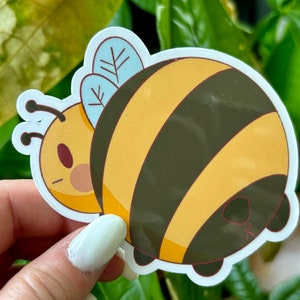Può includere: Un adesivo di ape di cartone animato giallo e nero con un'ala blu chiaro. L'ape è rotonda e ha un'espressione carina.