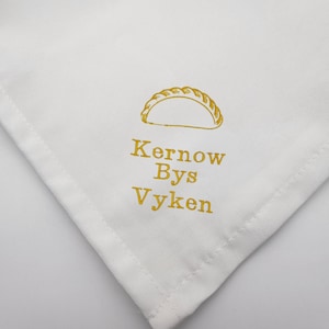 May include: White square cloth with a gold-coloured illustration of a Cornish pasty and the text "Kernow Bys Vyken". Green leaves are in the background.