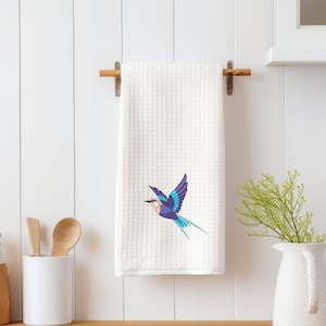 May include: White kitchen towel featuring a blue and purple bird design. The bird is depicted in flight with its wings outstretched. The towel is hanging on a wooden rack.