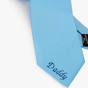 May include: A pale blue tie with a ribbed texture. The word "Daddy" is embroidered in a cursive font near the tie's pointed end. The tie is rolled up at the top, and the label reads "Hand Made Silk Dry Clean Only."