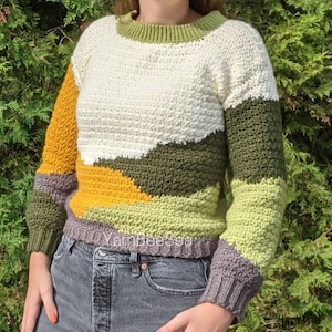 Könnte beinhalten: Ein gehäkelter Pullover mit einem mehrfarbigen geometrischen Muster in den Farben Weiß, Grün, Gelb und Grau.