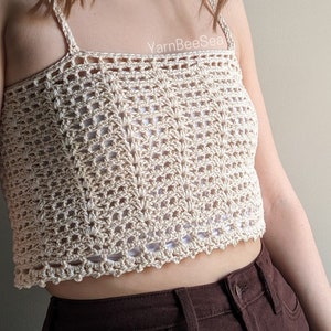 Könnte beinhalten: Ein gehäkeltes Crop-Top mit einem zarten, durchbrochenen Muster in hellbeige. Das Top hat dünne Träger und einen leicht verkürzten Saum.