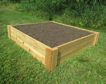 May include: A raised garden bed made of natural wood planks. The bed is filled with dark brown soil and is sitting on a green grassy lawn.