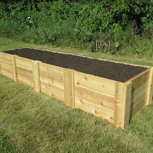 May include: A wooden raised garden bed with dark brown soil inside. The bed is made of natural cedar wood planks and is sitting on a green lawn.
