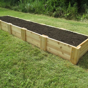 May include: A long, rectangular raised garden bed made of natural cedar wood. The bed is filled with dark brown soil and is sitting on a green lawn.