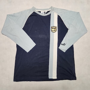 Può includere: Una maglietta blu navy a maniche lunghe con una striscia bianca e blu chiaro al centro. La maglietta ha una manica raglan blu chiaro e un piccolo logo Puma ricamato sulla manica sinistra. La maglietta ha anche un piccolo logo Puma ricamato con "48" all'interno sul lato destro del petto.