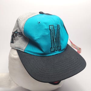 May include: A blue, grey and black baseball cap with a large black embroidered "M" on the front. The cap has a curved brim and a mesh back.