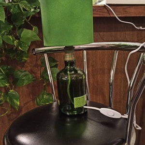 Pu&ograve; includere: Un paralume verde &egrave; posto sopra una bottiglia di vetro con un'etichetta che recita "Courvoisier" e un cavo bianco con un interruttore. La lampada &egrave; appoggiata su uno sgabello da bar nero con una base cromata.