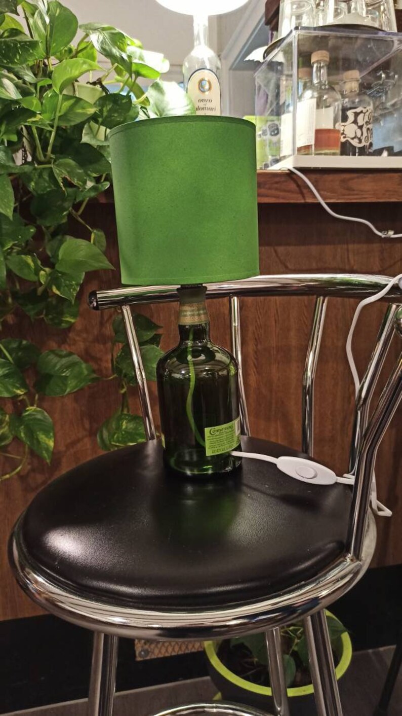 Pu&ograve; includere: Un paralume verde &egrave; posto sopra una bottiglia di vetro con un'etichetta che recita "Courvoisier" e un cavo bianco con un interruttore. La lampada &egrave; appoggiata su uno sgabello da bar nero con una base cromata.