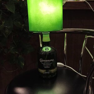 Pu&ograve; includere: Un paralume verde con una luce all'interno &egrave; posto su una bottiglia di vetro trasparente con un'etichetta verde su cui &egrave; scritto "Connemara Peated Single Malt Irish Whiskey Original". La bottiglia &egrave; appoggiata su uno sgabello da bar nero.