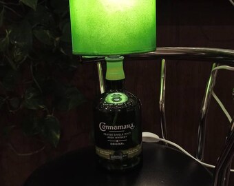 May include: A green lampshade with a light inside sits on top of a clear glass bottle with a green label that reads "Connemara Peated Single Malt Irish Whiskey Original". The bottle is sitting on a black bar stool.
