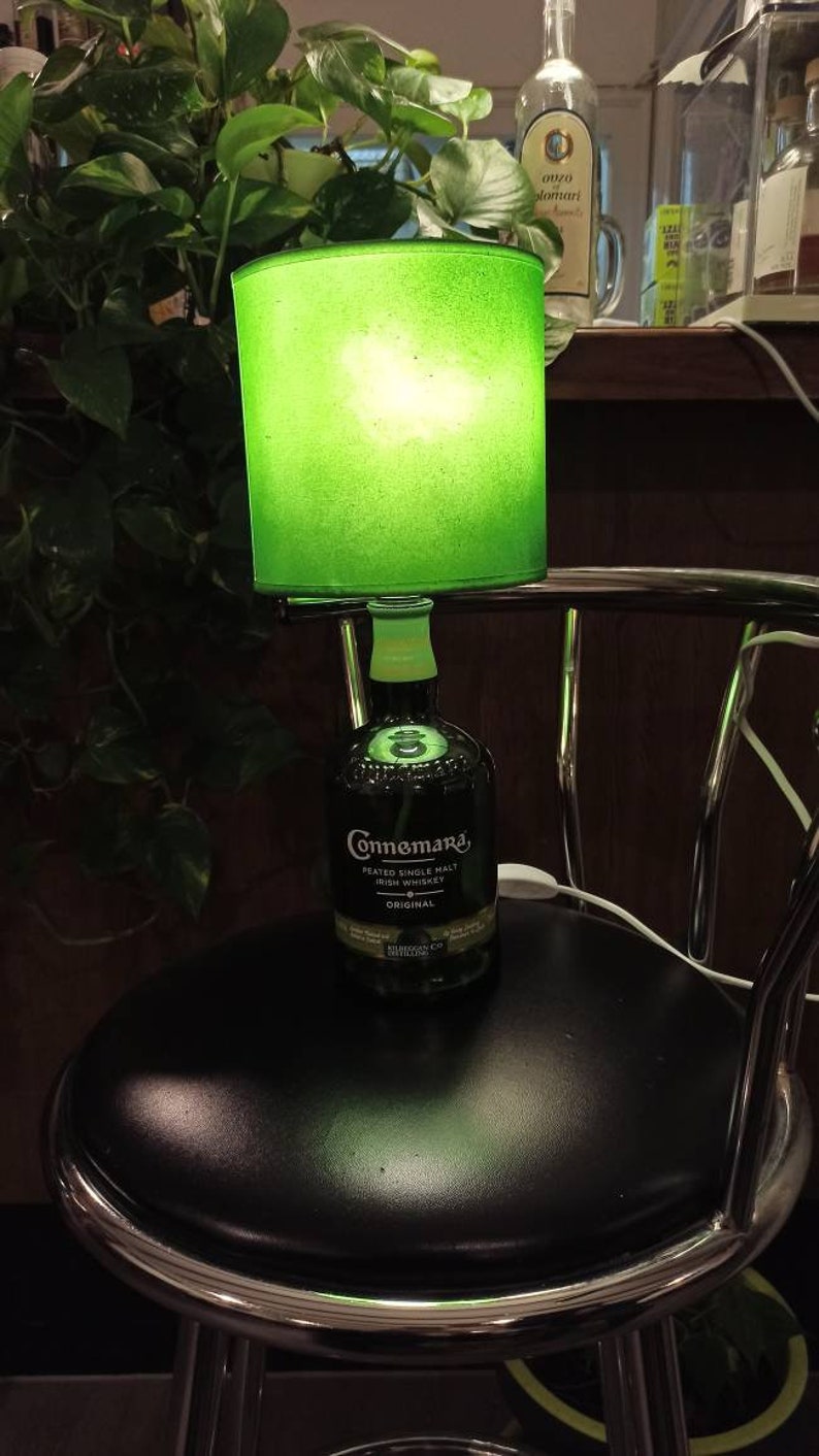 Pu&ograve; includere: Un paralume verde con una luce all'interno &egrave; posto su una bottiglia di vetro trasparente con un'etichetta verde su cui &egrave; scritto "Connemara Peated Single Malt Irish Whiskey Original". La bottiglia &egrave; appoggiata su uno sgabello da bar nero.
